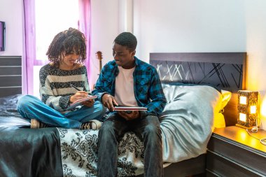 İki genç öğrenci bir yatakta oturuyor, derslerine çalışıyor, fikirlerini paylaşıyor ve rahat bir ev ortamında sınavlara hazırlanıyorlar.