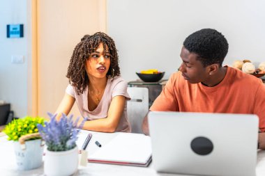 Evden çalışan çok ırklı genç bir çift, beyaz bir masada oturuyor, dizüstü bilgisayar kullanıyor ve tartışıyorlar.
