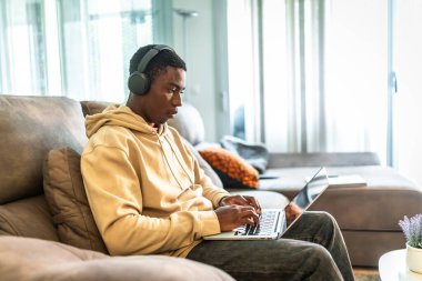 Genç adam rahat bir şekilde kanepede oturuyor, dizüstü bilgisayar ve kulaklık kullanıyor, modern bir oturma odasında evden çalışmaya ve çalışmaya odaklanıyor.