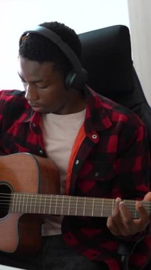 Genç Afrikalı Amerikalı müzisyen, ev stüdyosunda akustik gitar performansı kaydederken dikkatle odaklanıyor, kulaklık takıyor ve müzik yaratıcılığını ifade ediyor.