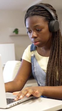 Derslerine odaklanan genç bir kız, ev ortamında kulaklık takarken dizüstü bilgisayarında daktilo kullanıyor.