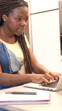 Genç öğrenci okul ödevi için özenle çalışıyor, parlak mutfakta dizüstü bilgisayar kullanıyor, araştırma projesini yazarken odaklanmış konsantrasyonu gösteriyor