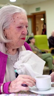 Yaşlı kadın bir masada oturuyor, bir bakımevinde bir fincan kahvenin tadını çıkarırken el kol hareketi yapıyor.