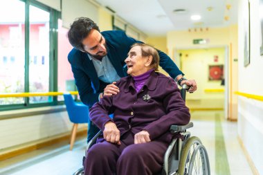 Doktor, yaşlı ve mutlu bir kadını tekerlekli sandalyeye itiyor. Huzurevinin koridorunda gülümsüyor ve onunla konuşuyor.