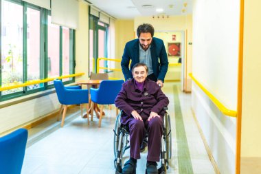 Erkek hemşire, huzurevinin koridorunda tekerlekli sandalyedeki mutlu yaşlı kadına yardım ediyor.