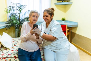 Hemşire, huzurevinde akıllı telefon kullanan yaşlı bir kadına yardım ediyor. Teknolojik destek ve rehberlik sağlıyor.