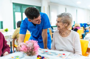Genç erkek hemşire, bir bakımevinin ortak odasında, gülümseyen yaşlı bir kadına masa oyunu oynaması için yardım ediyor, bilişsel sağlık ve sosyal etkileşimi teşvik ediyor.