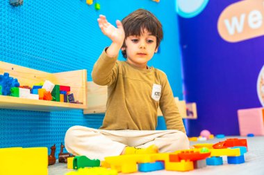 Yerde oturan genç çocuk, renkli inşaat oyuncaklarıyla meşgul ve hareketli bir anaokulunda yapı taşları yapıyor.