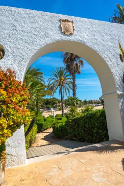 Beyaz bir kemer yolu boyunca manzara, açık mavi gökyüzünün altında yemyeşil ve palmiye ağaçlarıyla çevrili güzel Roc de Sant Gaieta plajına giden yolu gösteriyor.