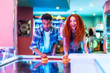 Hava hokeyi oynayan iki genç arkadaş, renkli bir oyun salonunda birlikte hafta sonunun tadını çıkarıyorlar.