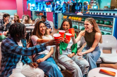 Bir grup neşeli, etnik çeşitliliğe sahip genç arkadaş, bovling salonunda eğlenceli bir hafta sonu geçirirken kırmızı plastik bardaklarla kadeh kaldırıyor.