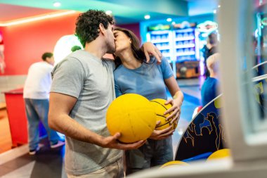 Romantik çift, arkadaşları ve atari oyunlarıyla çevrili canlı bir eğlence merkezinde basketbol ve bowlingin keyfini çıkarırken öpüşüyor.