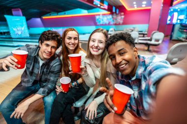 Bir grup neşeli, etnik çeşitlilikte genç arkadaş içki içip bowling oynarken selfie çekiyorlar.