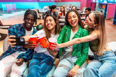 Çok ırklı dört genç arkadaş kadeh kaldırıyor ve bowling salonunda selfie çekiyorlar. Birlikte hafta sonunun tadını çıkarıyorlar.