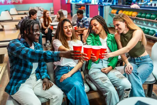 Bir grup neşeli, etnik çeşitliliğe sahip genç arkadaş, bowling salonunda eğlenceli bir hafta sonu geçirirken kırmızı bardaklarla kadeh kaldırıyor.