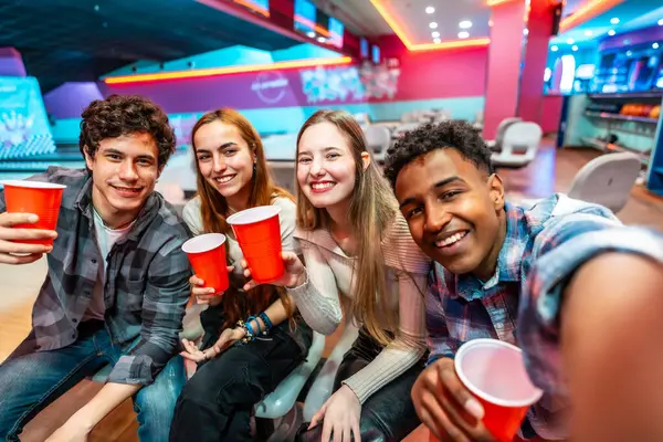 Bir grup neşeli, etnik çeşitliliğe sahip genç arkadaş bir bowling kulübünde içerken ve eğlenirken selfie çekiyorlar.