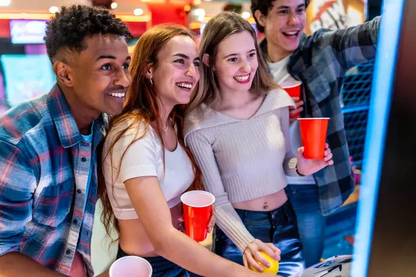 Neşeli, çoklu etnik kökenli bir grup arkadaş birlikte eğleniyor, atari oyunları oynuyor ve bowling salonunda kırmızı bardaklar tutuyor.