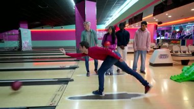 Bir grup arkadaş bowling oynarken, bir genç adam top atmaya hazırlanırken diğerleri ona tezahürat yapıyor.