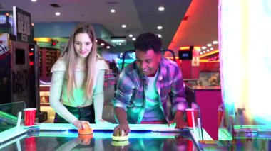 Çok ırklı genç arkadaşlar, neon ışıklarıyla renkli atari salonunda oynarken gülüp kutlama yaparak, rekabetçi hava hokeyi maçının keyfini çıkarıyorlar.