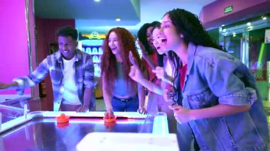 Genç arkadaşlar canlı atari salonunda hokey oynarken yarışıyor ve gülüyorlar, enerjik grup dinamikleri ve gençlik keyfi sergiliyorlar.