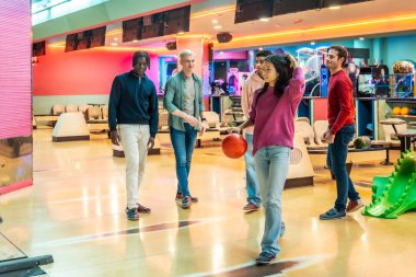 Genç bir kadın, neşeli bowling salonunda tezahürat yapan arkadaşları tarafından çevrelenmiş bir bovling topunu sokağa atıyor.