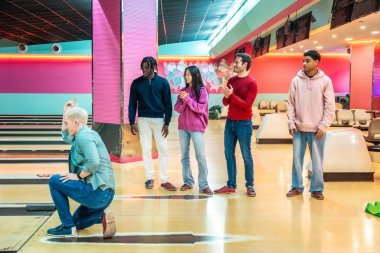 Çeşitli genç arkadaşlar birlikte bovling oynarken eğleniyorlar, biri topu fırlatırken diğerleri tezahürat yapıyor ve alkışlıyor.