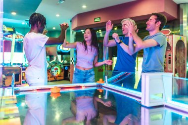 Çoklu etnik arkadaşlar beşlik çakıyor, zaferlerini canlı bir oyun salonunda hava hokeyi masasında kutluyor.