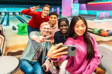 Bir grup neşeli, etnik çeşitliliğe sahip genç arkadaş akıllı telefonlarıyla selfie çekiyor ve bowling salonunda birlikte vakit geçirirken barış işareti yapıyor.