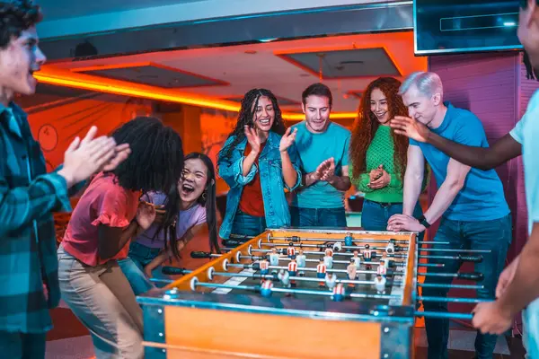 Çok ırklı arkadaşlar langırt oynuyorlar, alkışlıyorlar ve gülüyorlar, canlı bir eğlence mekanında.