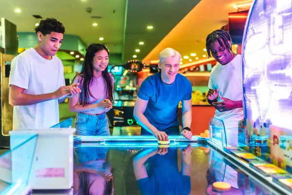 Cok etnik arkadaslar renkli isiklarla dolu, eglenceli bir atari salonunda hokey oynayip gülüyorlar.