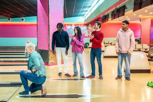 Çeşitli genç arkadaşlar birlikte bovling oynarken eğleniyorlar, biri topu fırlatırken diğerleri tezahürat yapıyor ve alkışlıyor.
