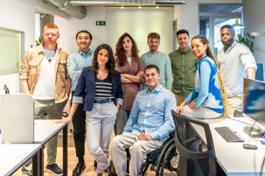 Modern bir ofiste birlikte duran çok kültürlü profesyonellerden oluşan bir ekip, tekerlekli sandalye kullanan bir meslektaşla çeşitlilik ve kapsama sergilediler.