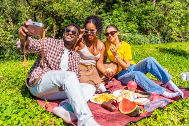 Üç arkadaş, parkta piknik yaparken eski bir kamerayla selfie çekiyorlar.