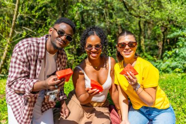 Üç arkadaş, bereketli bir parkta yaz pikniğinde karpuz dilimleri paylaşıyor güneşli havanın ve birbirlerinin arkadaşlığının tadını çıkarıyorlar.