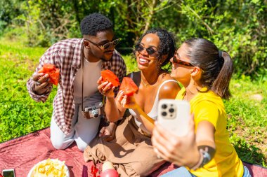 Üç arkadaş parkta piknik yapıyorlar, karpuz yiyorlar ve akıllı telefonla selfie çekiyorlar.