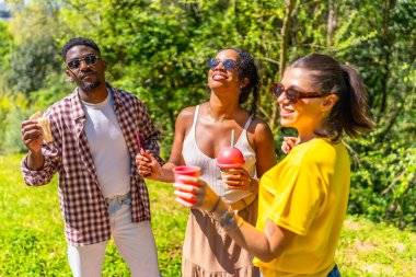Üç neşeli genç arkadaş bereketli yeşilliklerle çevrili canlı bir yaz festivalinde ferahlatıcı içecek ve atıştırmalıkların tadını çıkarıyorlar.