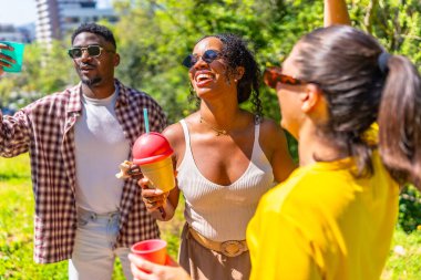 Neşeli bir grup arkadaş güler, dans eder ve canlı bir park ortamında içkilerin keyfini çıkarırlar.