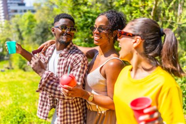 Üç arkadaş güneşli bir günde parkta yürüyor, içkinin ve arkadaşlığın tadını çıkarıyorlar.