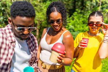 Üç arkadaş güneşli bir parkta birlikte ferahlatıcı içecekler içiyorlar. Arkadaşlıklarının ve güzel havanın tadını çıkarıyorlar.