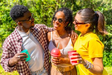 Üç genç çok ırklı arkadaş güneşli bir günün tadını çıkarıyor, içkilerini yudumluyor ve gülümsüyor.