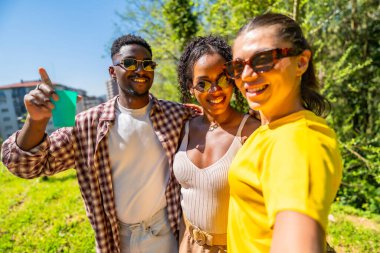 Güneş gözlüğü takan üç mutlu çoklu etnik arkadaş parkta fotoğraf çekiyor, kameraya bakıyor ve gülümsüyor.