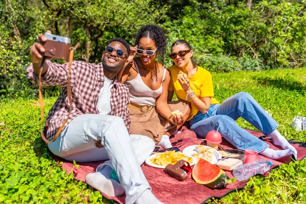 Üç arkadaş, parkta piknik yaparken eski bir kamerayla selfie çekiyorlar.