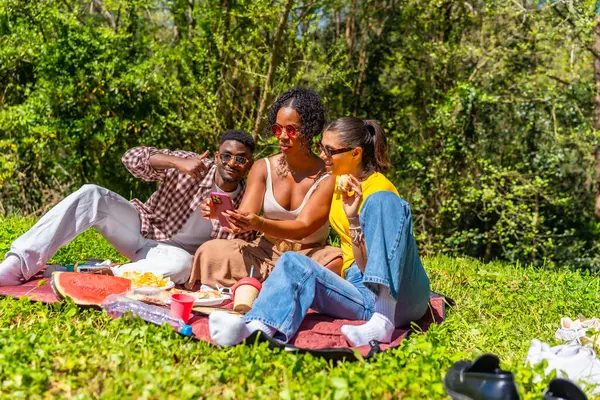 Üç arkadaş parkta piknik yapıyor, selfie çekiyor ve birlikte vakit geçiriyorlar.