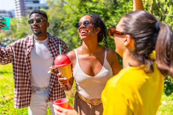 Neşeli bir grup arkadaş güler, dans eder ve canlı bir park ortamında içkilerin keyfini çıkarırlar.