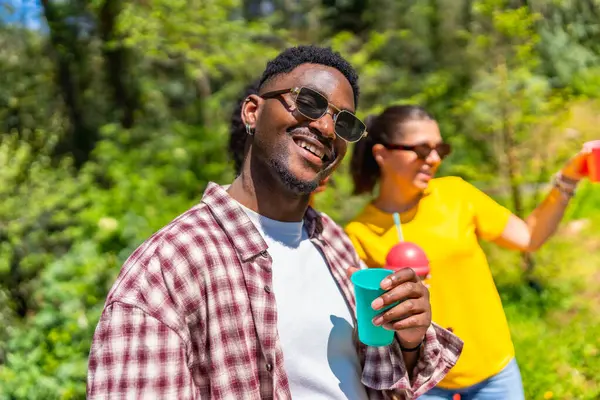 Güneşli bir günde gülümseyen ve ferahlatıcı bir içki tutan genç adam, doğal ortamda arkadaşlarıyla eğleniyor.