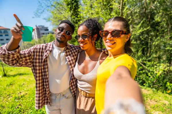 Parkta selfie çeken üç mutlu etnik arkadaş. Bir tanesi parmağını gökyüzüne doğrultuyor.