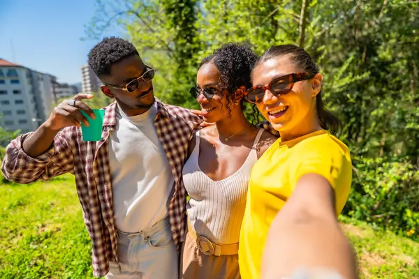Üç neşeli çoklu etnik arkadaş parkta selfie çekiyorlar, güneşli bir günün tadını çıkarıyorlar.
