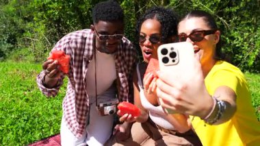 Üç arkadaş piknik yapıyorlar, karpuz dilimleri yiyorlar ve akıllı telefonla selfie çekiyorlar.