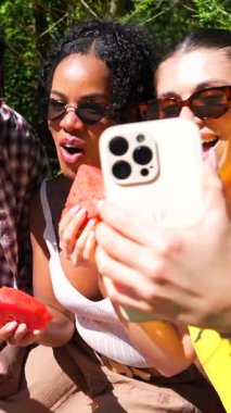 Bir grup arkadaş akıllı telefonla fotoğraf çekiyor ve parkta piknik yaparken karpuz dilimleri yiyorlar.