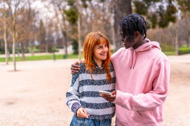 Neşeli bir parkta akıllı bir telefona bakarken gülümseyen, güneşli bir günde birbirlerinin arkadaşlığından keyif alan çok ırklı genç bir çift.